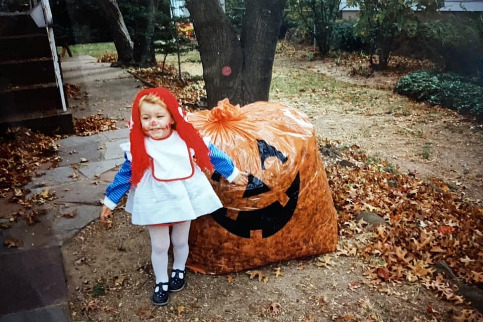 A photo of the author as a toddler dressed up as Raggedy Ann, standing in front of a large pumpkin leaf bag.