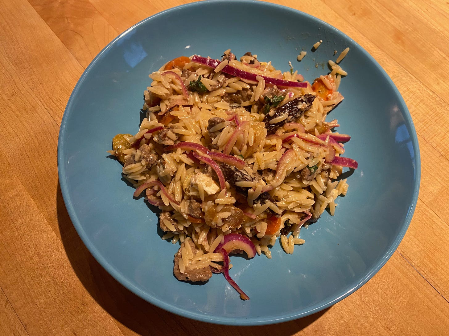 A pile of orzo, slices of lamb, dates, and slivers of pickled red onion in a shallow blue bowl on a kitchen counter.