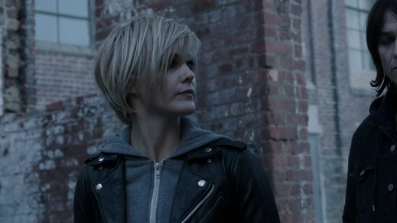 Elizabeth standing outside wearing a short blond wig, grey sweatshirt, and black leather jacket.