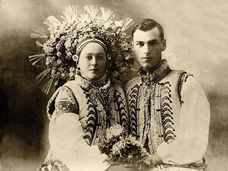 A lovely young couple in ornate traditional dress stare proudly into the camera. The bride is wearing a gorgeous floral wreath.