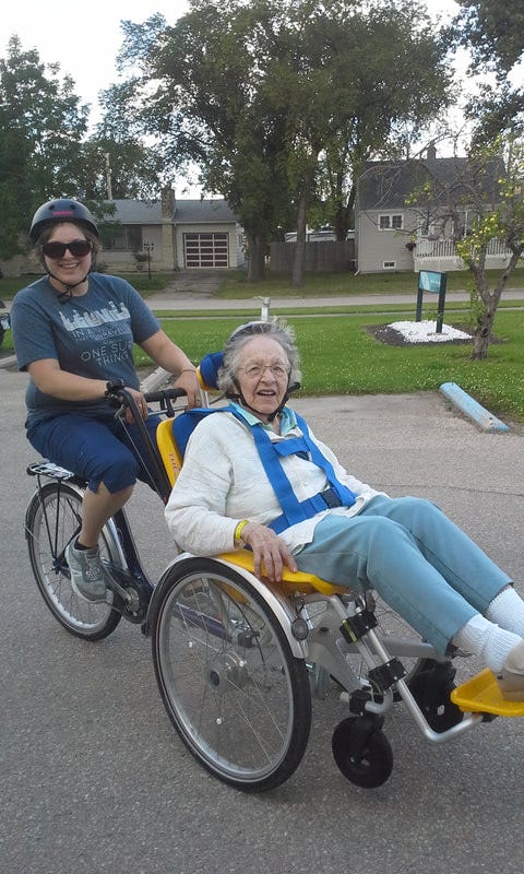 A young woman riding a bicycle with an additional seat in front, where an older woman is sitting, smiling as they ride past houses on a street.