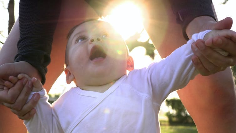 A toddler tries to walk outdoors, with an adult guiding it by both hands