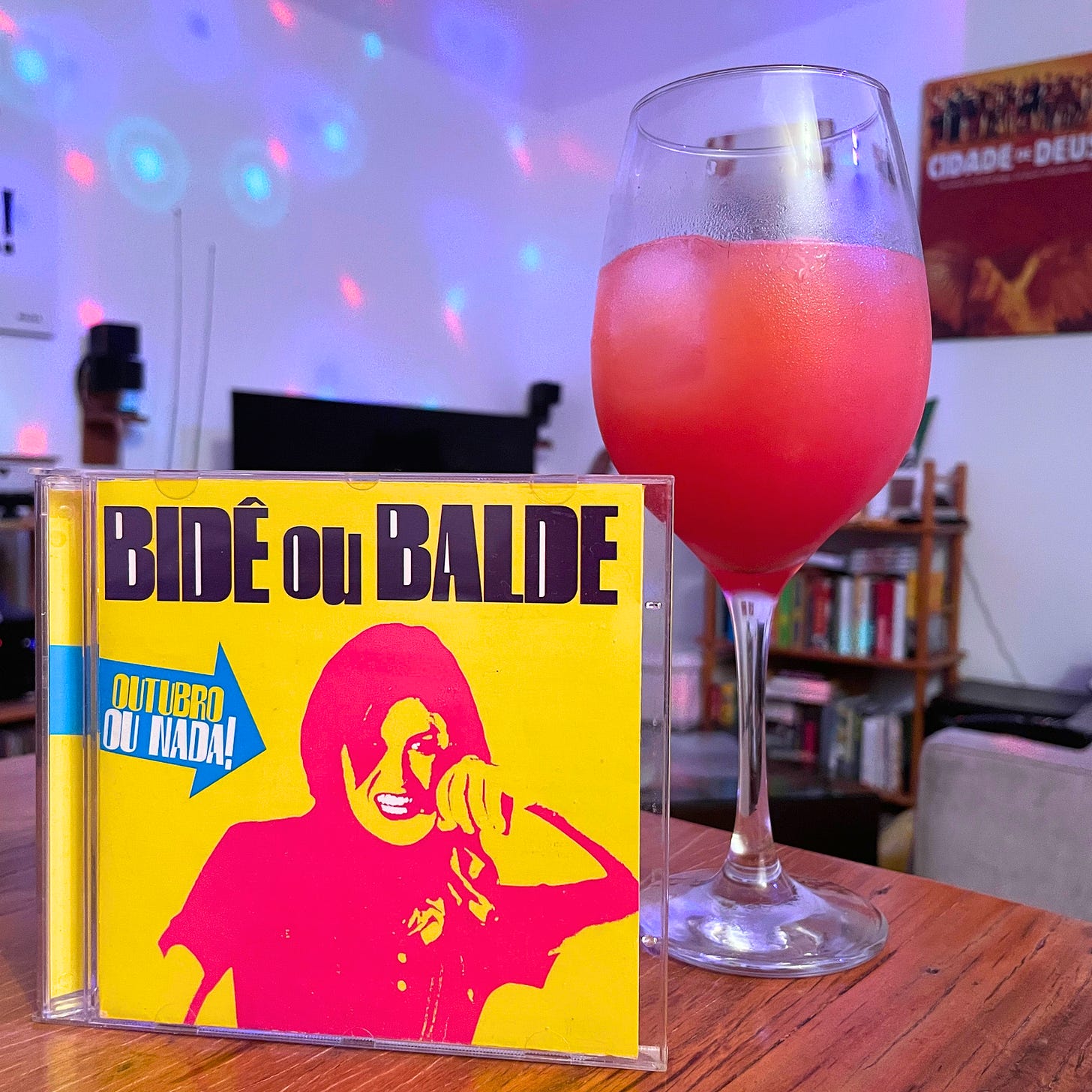 Texto mostra o disco Outubro ou Nada, de fundo amarelo e uma moça em grafismos rosas, uma taça de vinho com o drink Garibaldi, e ao fundo, uma sala de estar bagunçada