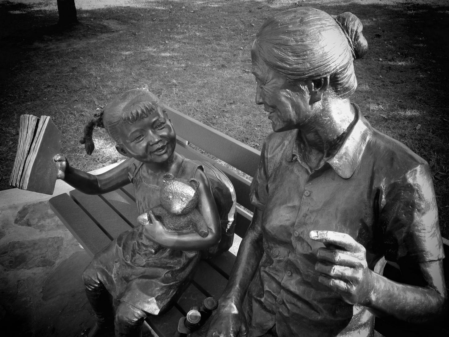 Photograph of bronze statue of grandmother and little girl on a park bench, looking at each other and smiling. The little girl is holding a book up. Statue in Sugar Land, Texas.