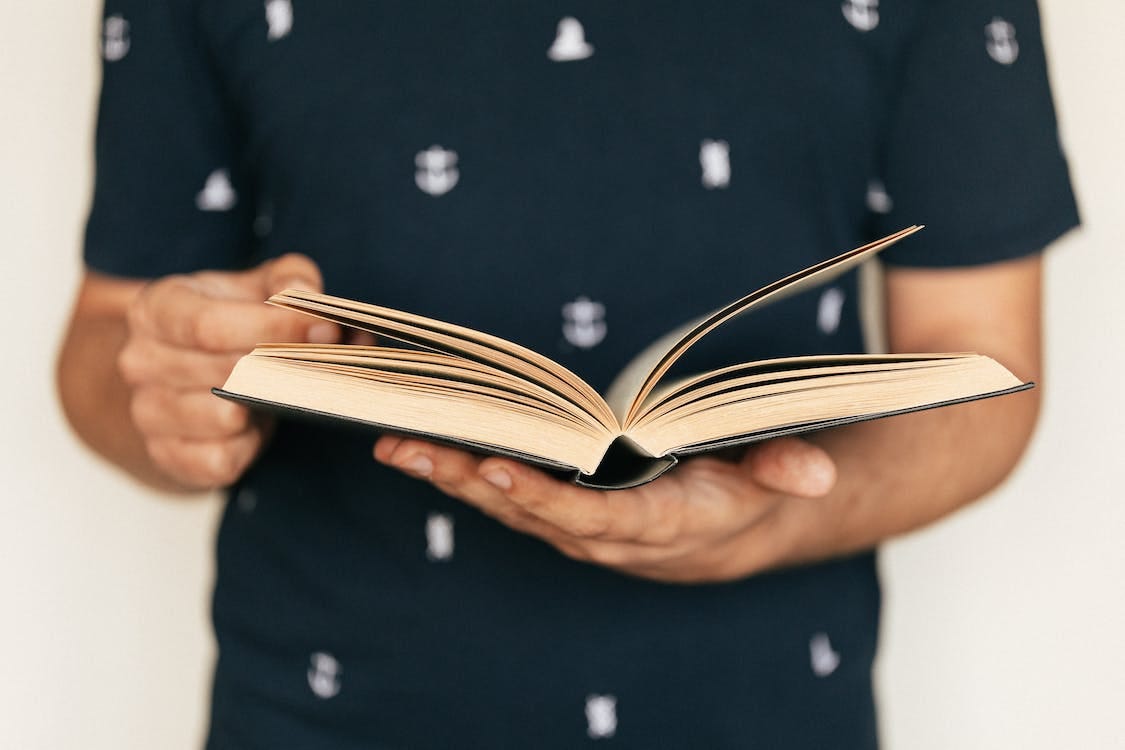 Free Anonymous male in dark tee shirt reading interesting book while standing in front of light wall Stock Photo