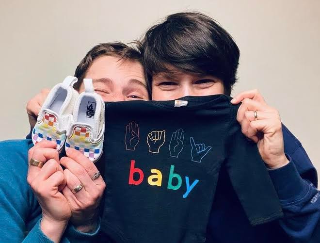 Two parents peeking behind a baby onesie that reads in sign language "baby" and lettering "baby" and a pair of white rainbow vans shoes