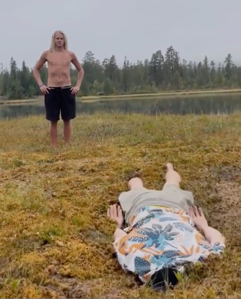 A blonde, shirtless man stands in a bog, against a gray sky and nondescript forest. In front of him another man lies face down in the bog wearing a floral shirt.