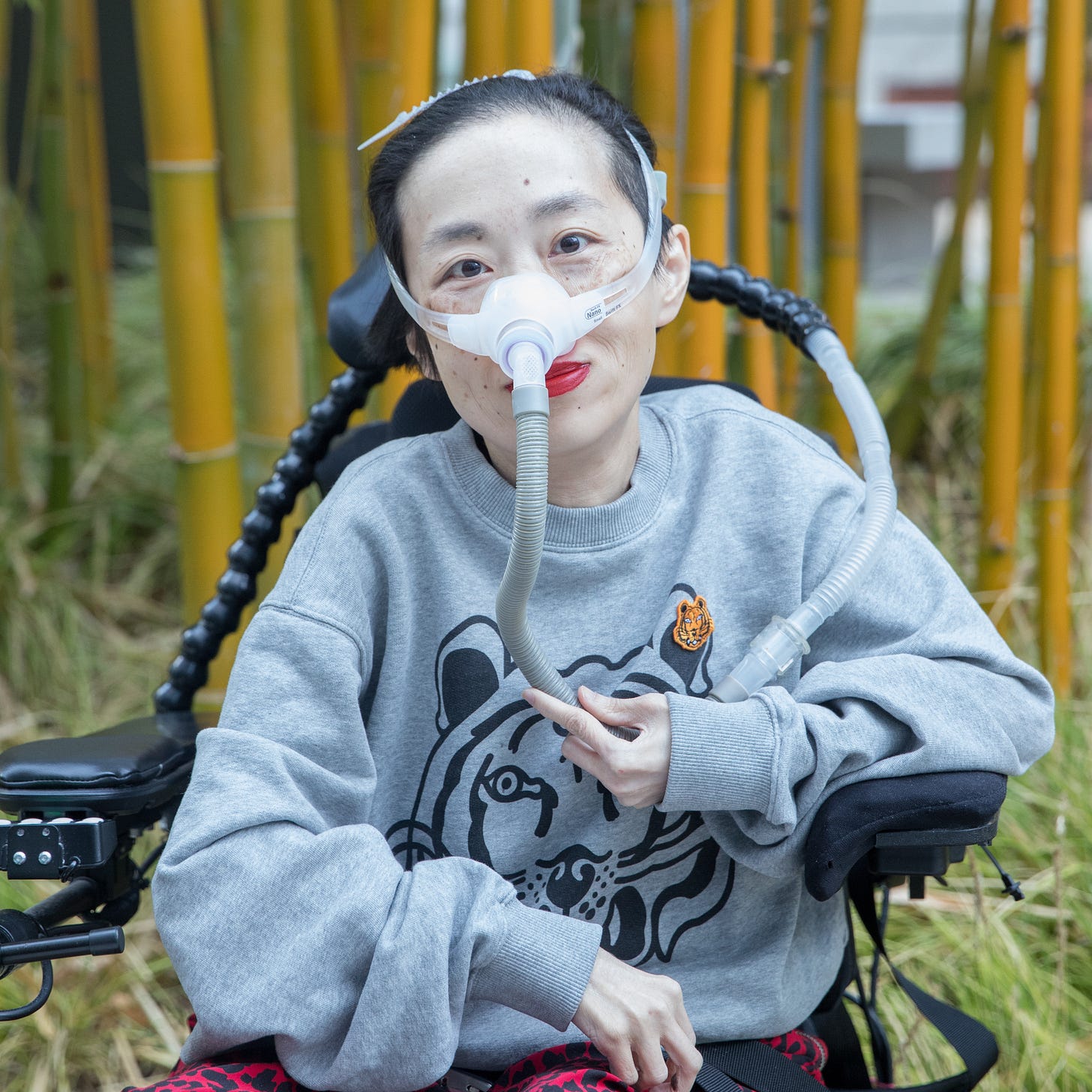 Photo of Alice Wong, an Asian American disabled woman with a mask over her nose attached to a tube for her ventilator. She is in a power wheelchair and wearing a gray sweatshirt with a tiger and leopard-print red and black pants. Behind her are bamboo trees. Credit: Eddie Hernandez Photography