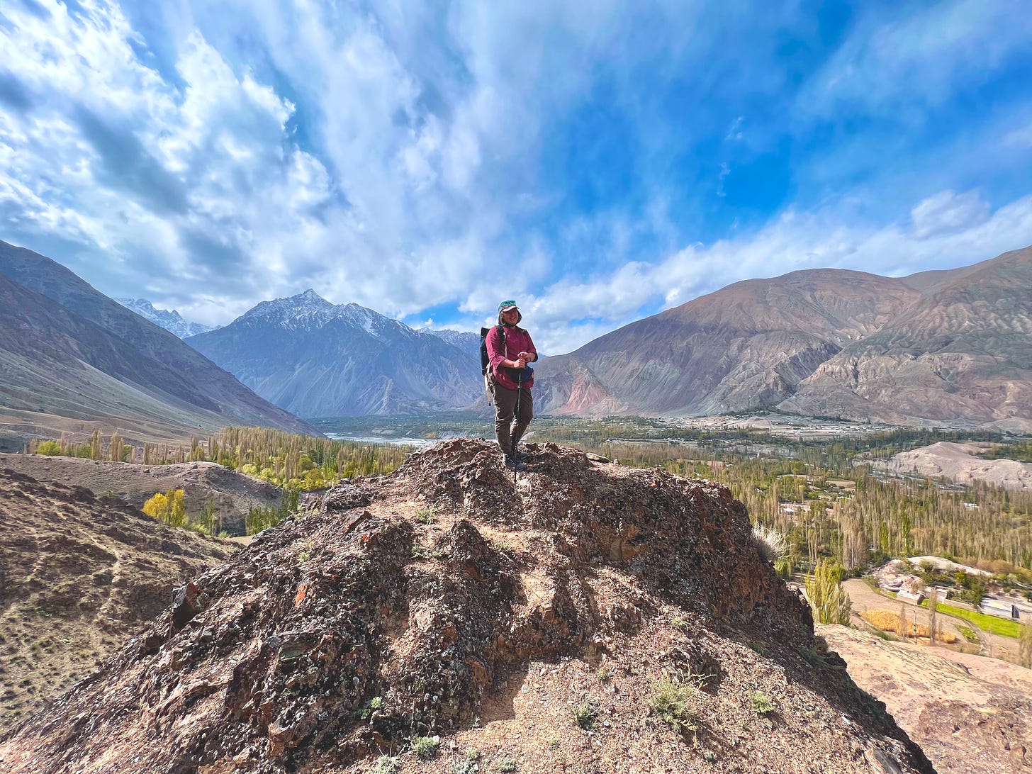 Asian girl standing on top of a rock on top of a hill in Yasin Valley, Pakistan. Behind her is the valley with green fields, blue river, green trees, and a large brown mountain range