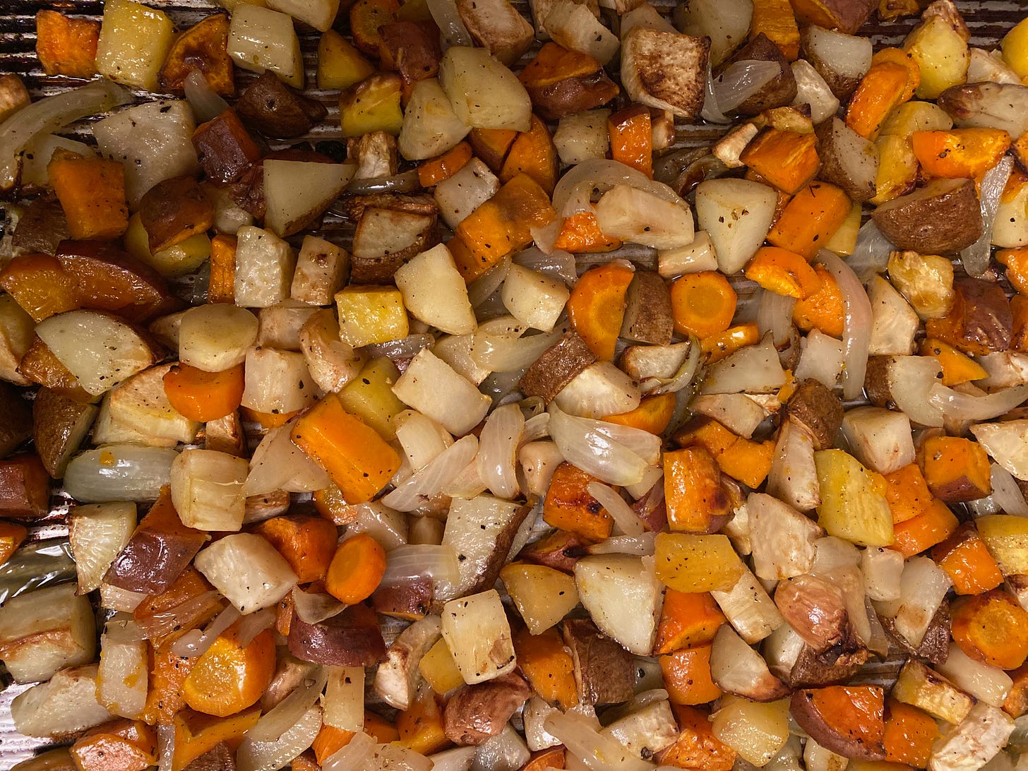Closeup of a pan of roasted roots—carrots, turnips, potatoes, parsnips, sweet potatoes, wedges of onion, and whole garlic cloves.