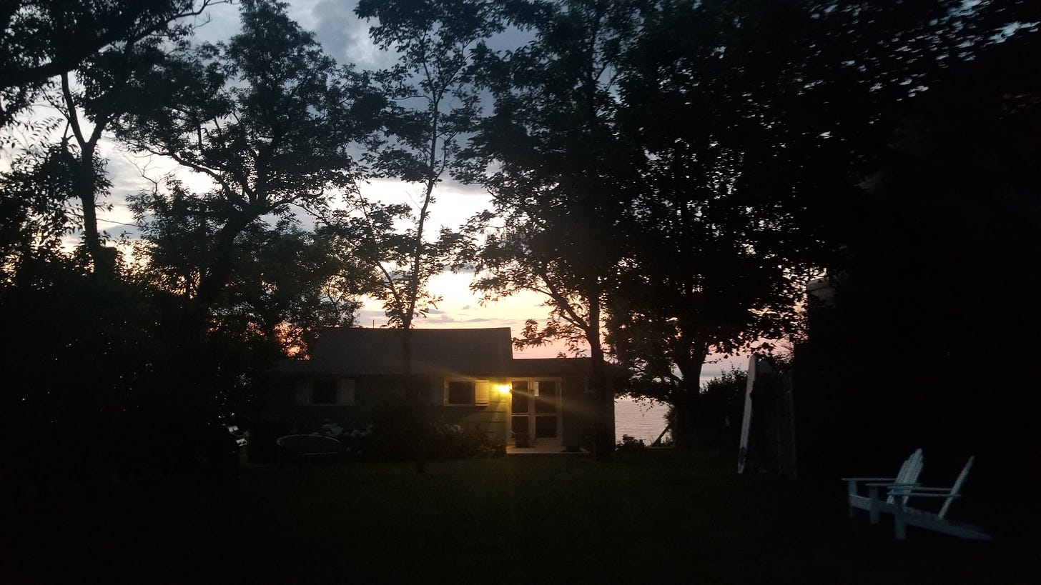 A tiny cottage stands before a fading sunset. You can make out the open water beyond. A single light shines on the back deck.
