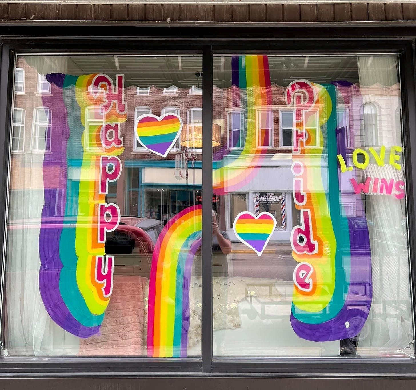 A storefront window is decorated with rainbow hearts and strips, the words "Happy Pride" and "Love Wins" in bright pink and yellow lettering.