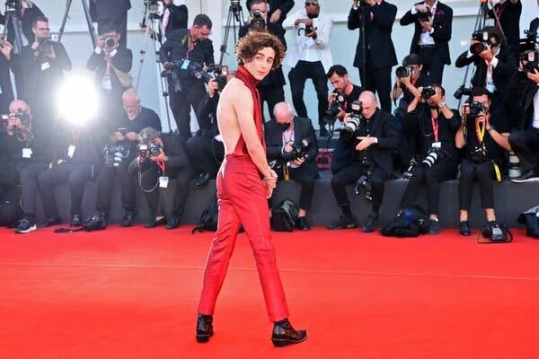 Timothée Chalamet, wearing a red halter top, red pants and black shoes, looking over his shoulder on the red carpet at the Venice Film Festival.
