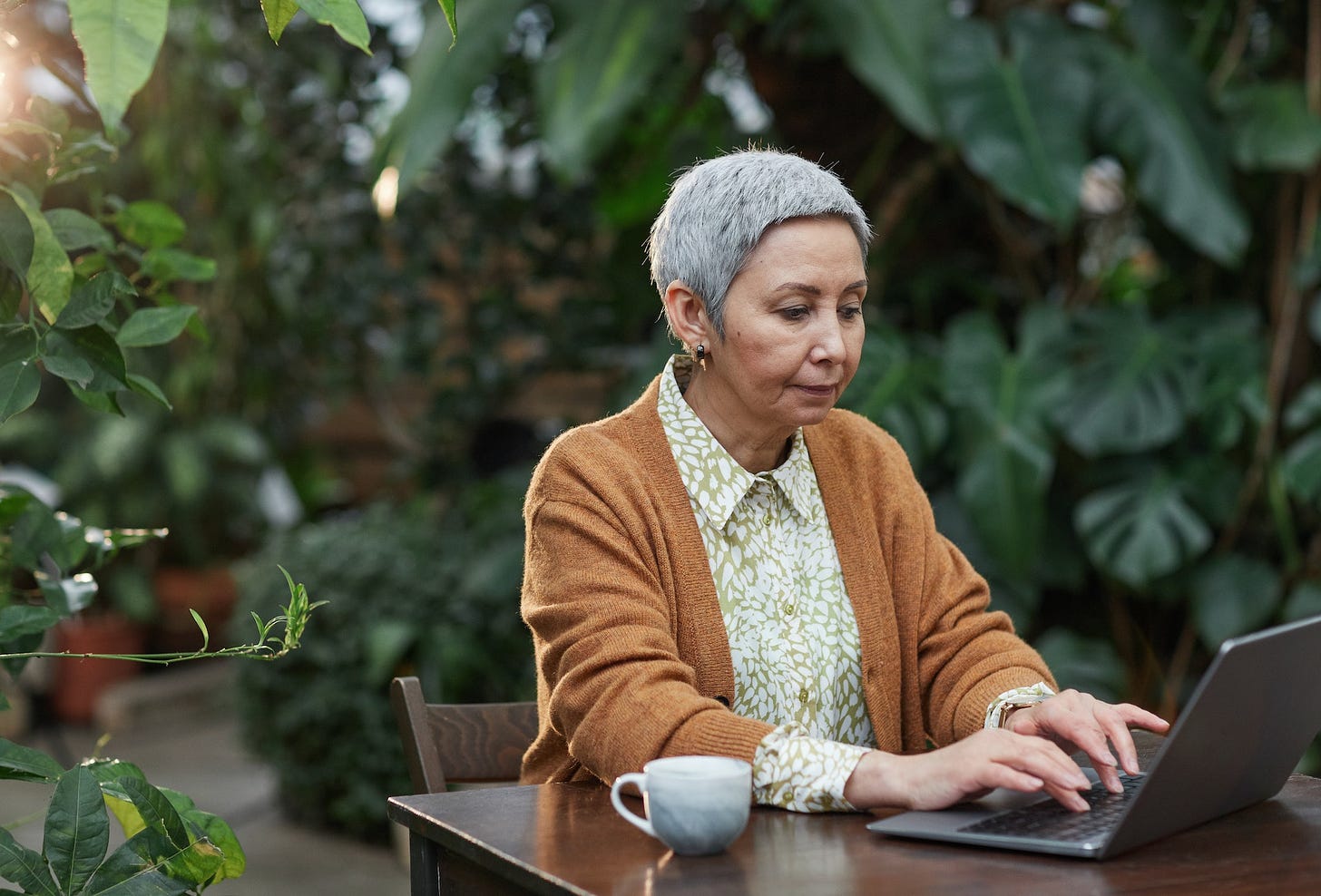 Elderly woman with short grey hair using an Apple Macbook outside with a white mug next to her. The elderly woman is wearing a floral top and a brown jacket and is sat in a leafy scene outside her home.