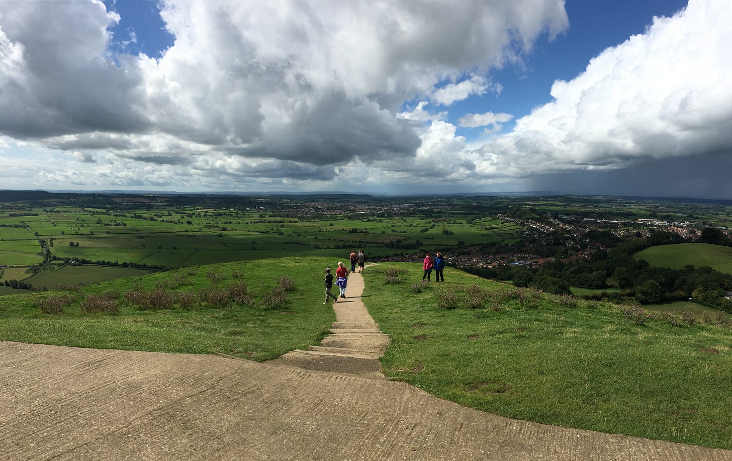 Partly cloudy sky over a vast expanse of green. A dirt path cuts down a hill in the center of the image, toward towns and houses below. A few people are ahead on the path.