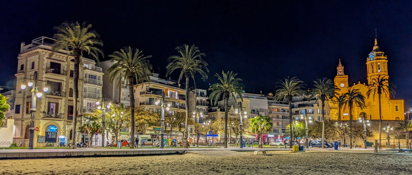 A picture of Sitges at night as seen from the beach. Tall palm trees stand in front of hotels lining the beach. 