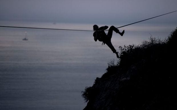 Silhouette of a tightroope walker with the ocean behind him. He's fallen off the rope, but managed to hold on.