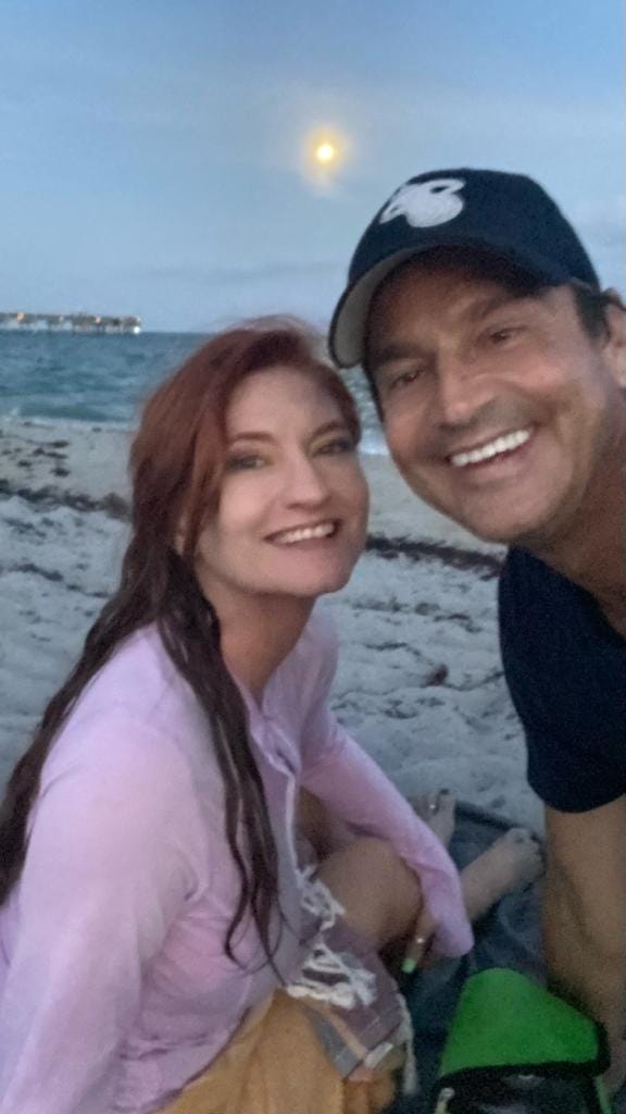 Two smiling white people on a beach at twilight with a blurry moon and pier in the background.