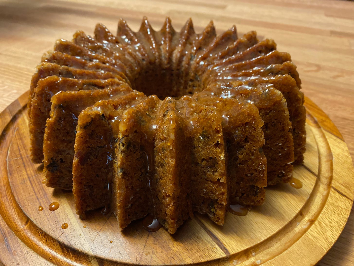Close-up of a golden brown tea cake sitting on a wooden plater. It’s a round cake with a hole in the center, with pronounced ridges from the patterned bundt pan it was baked in. A glaze shimmers on top. 