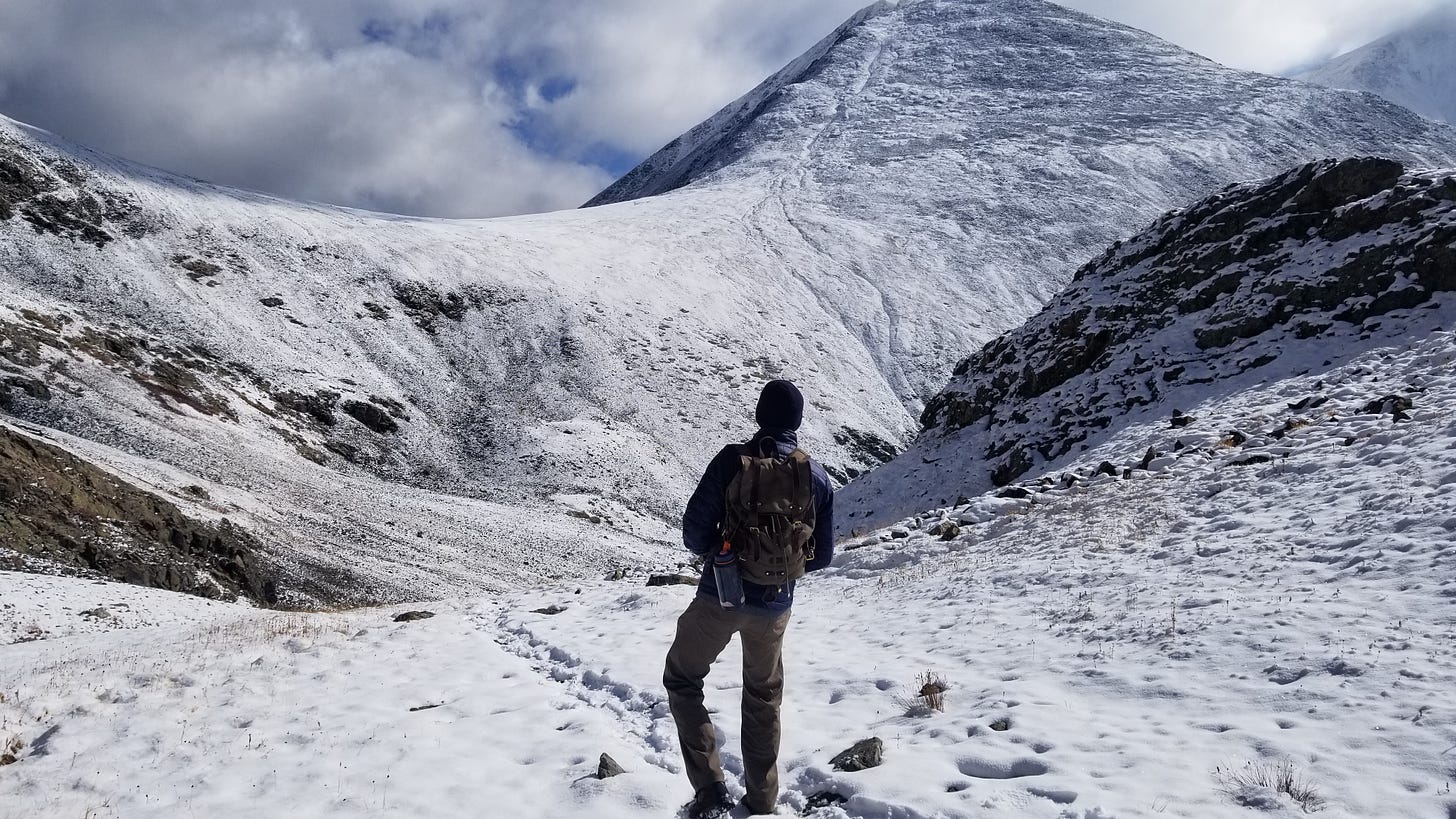 A hiker looks up at the snow-capped ridgeline. Clouds fill the sky. Snow covers the ground.