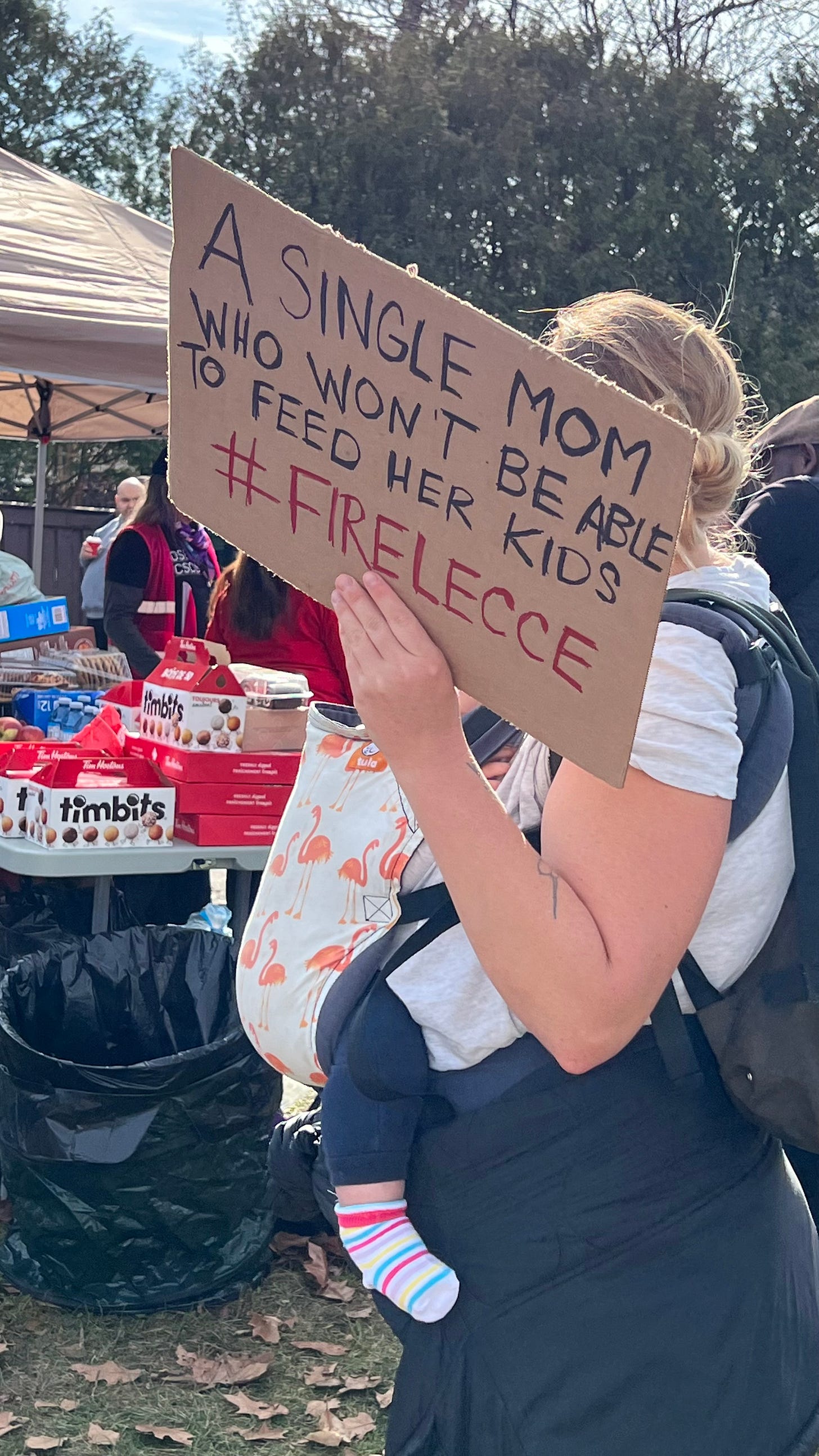 A woman carrying a child holds a sign that says "Single mom who won't be able to feed her kids #FIRELECCE