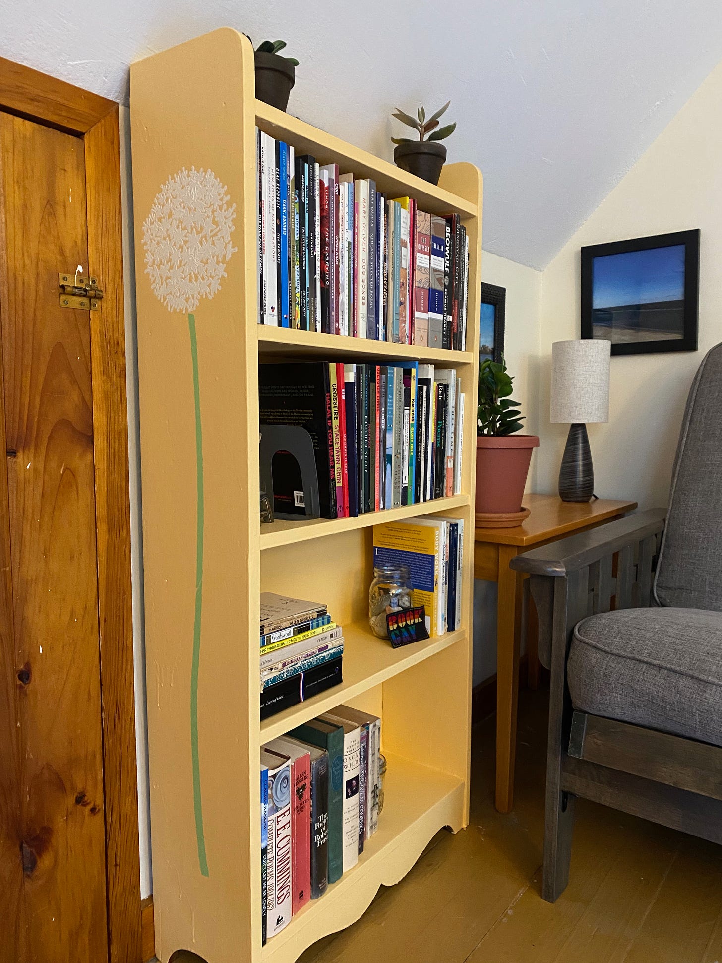 A small yellowy-orange bookshelf full of books and plants in the corner of a room. A large allium flower, with a green stem and white head, is stenciled onto the side of the bookshelf.