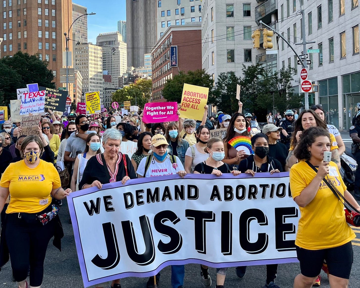 A large group of people march through the streets of New York City. In front, several people carry a sign that reads, "We demand abortion justice."