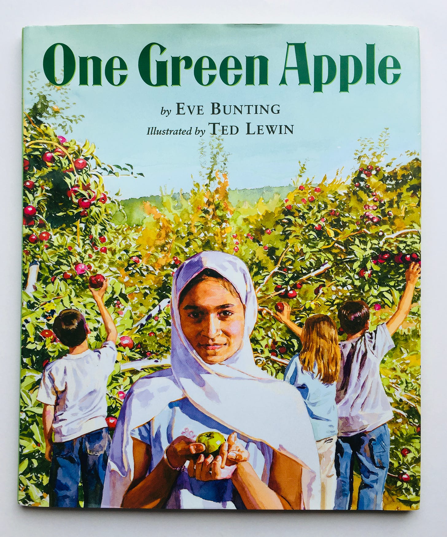 A girl with her head covered holds up a green apple in front of other children picking apples from trees