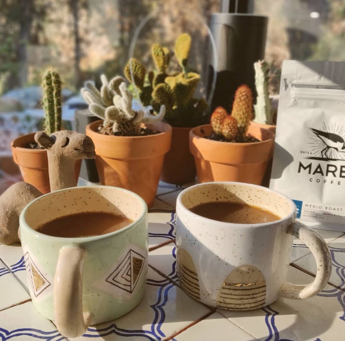 Two clay mugs of coffee on a white tile table outside with red clay pots with succulents and a bag of marea coffee in the background.