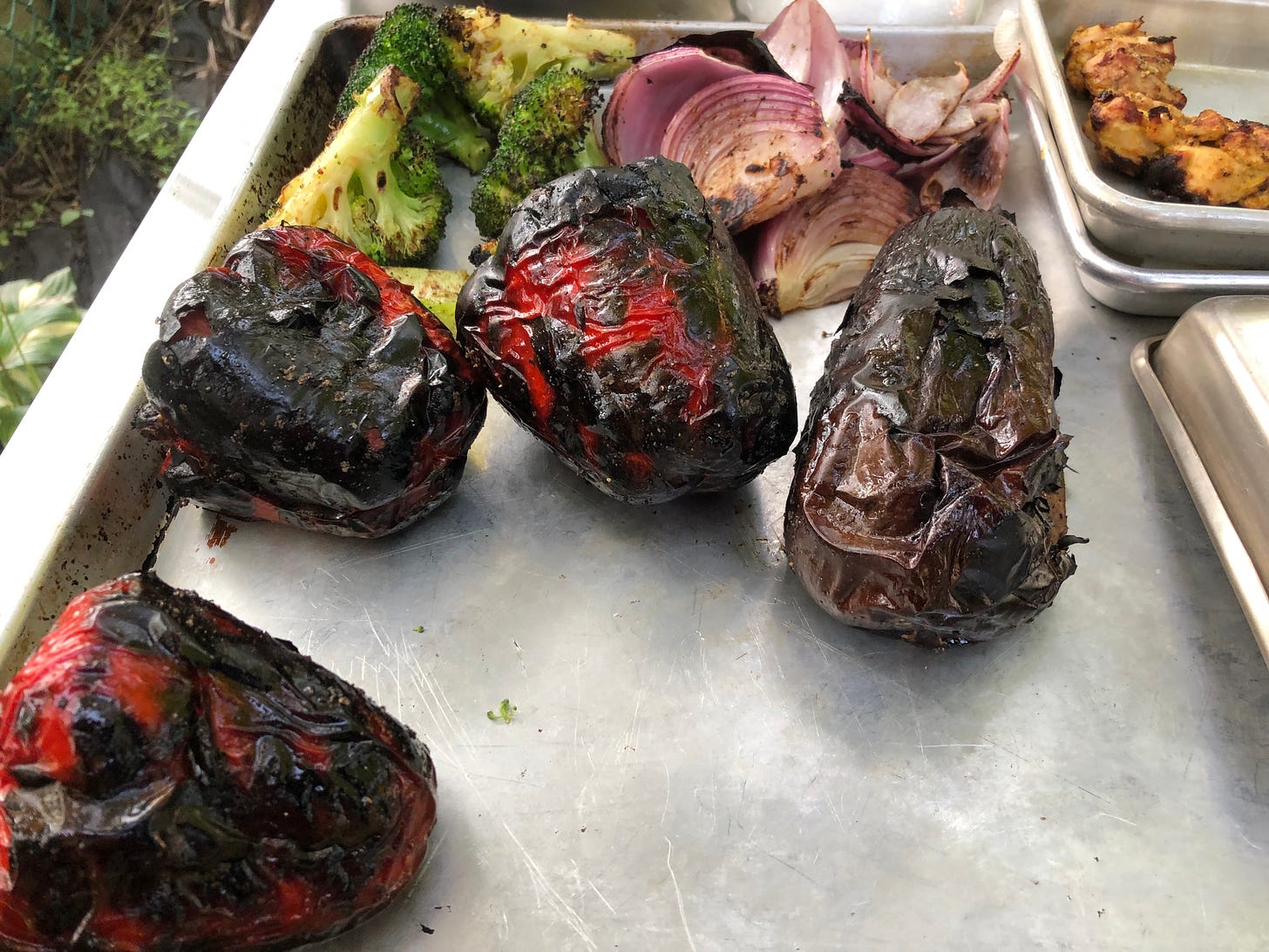 A tray holding grilled broccoli chunks and grilled red onion in the background. In the foreground, there are three whole grilled red peppers and a whole grilled eggplant, all with their skin very charred. 