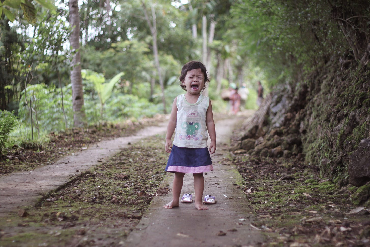 A young child barefoot on a path wearing a shirt that says 'happy' while open-mouthed and obviously screaming mid-meltdown.