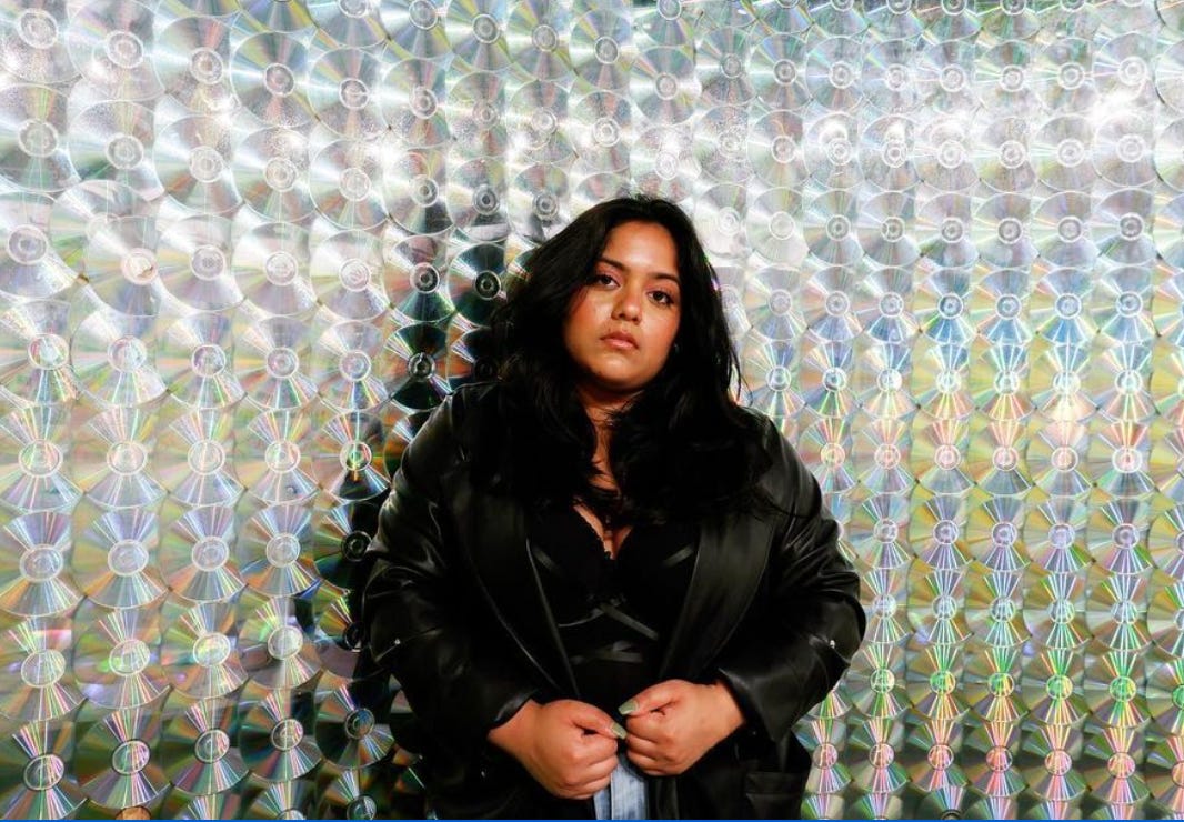 renuka standing in front of a wall of CD's wearing a leather jacket