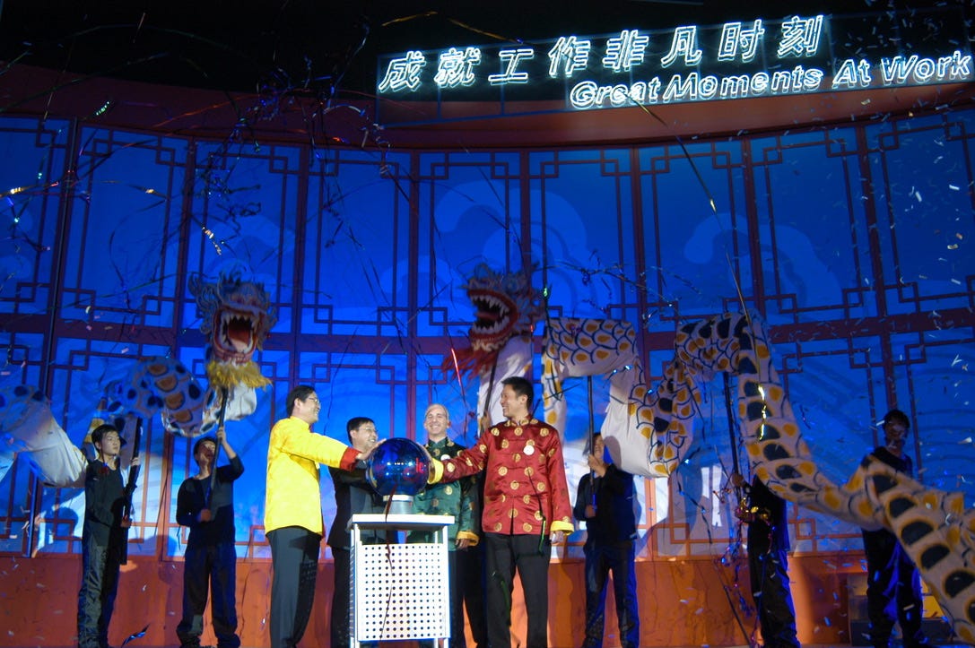 A very festive stage with "Great Moments At Work" in English and mandarin neon at the top, paper dragons, and a lighted ball that each of us are touching. We are wearing traditional chinese celebration garments.