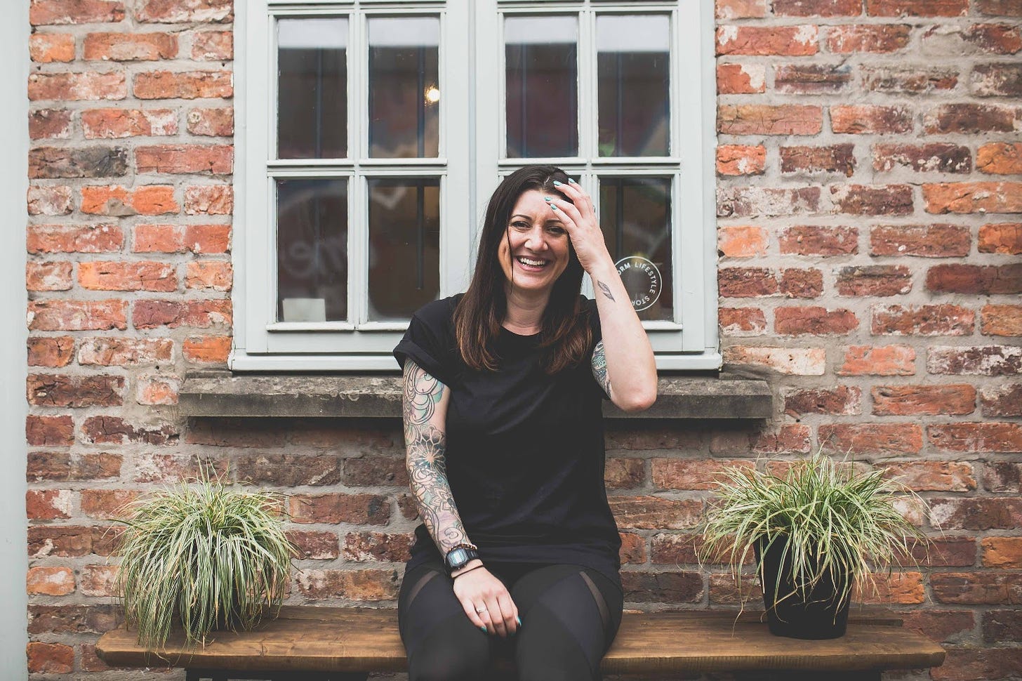 White woman in her 30s in a black T-shirt and jeans sits on a bench outside a redbrick house, under a window. Two plants sit either side. She's laughing and adjusting her hair with one hand.