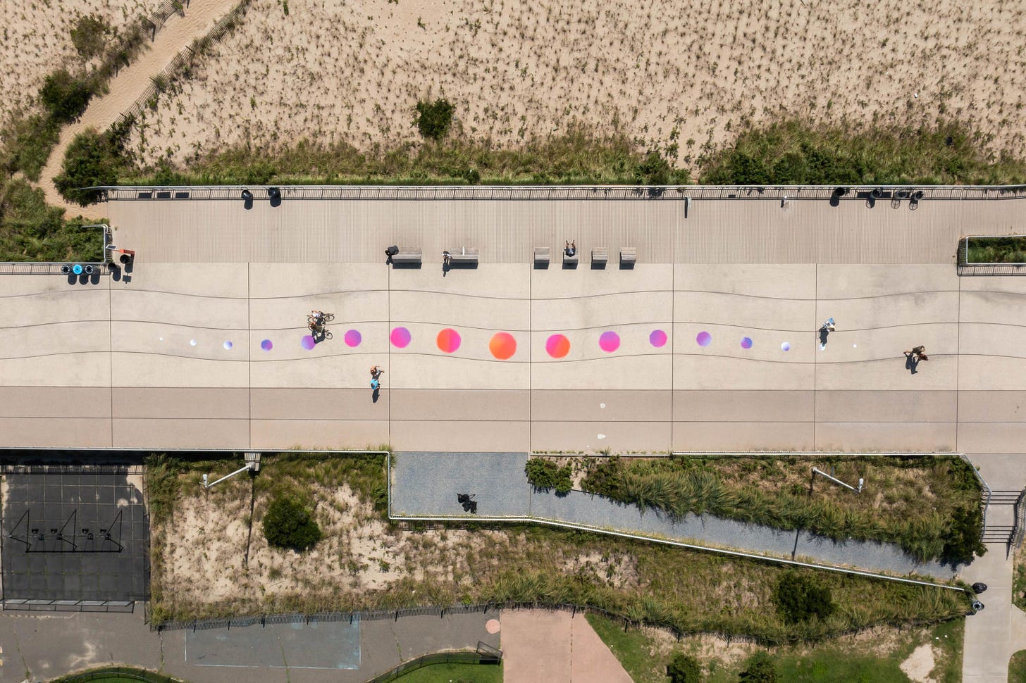 A shot from above looking straight down at the ground, like you’re within 5 minutes of landing on an airplane. Along a cement boardwalk next to a sandy beach, there are colorful spots that grow larger in sequence, gently curving within the wavy lines of the cement blocks.