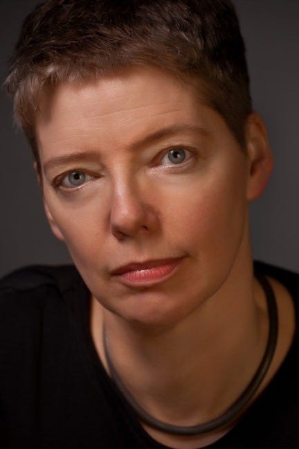 Photo of a Nicola Griffith, white woman with short blonde hair. She is staring intently at the camera. The background behind her is completely black. She has a black scoop neck shirt and a metal necklace.