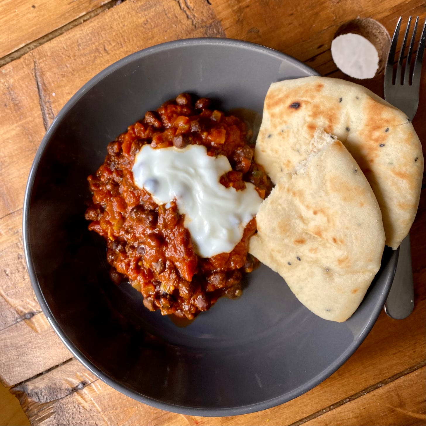A grey bowl on a wooden table, with chickpea masala topped with a dollop of yoghurt. Toasted naan bread to the side.