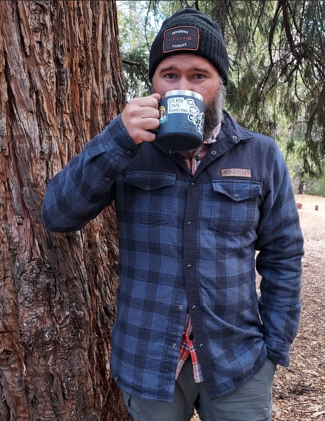 Bearded, white guy, with a thick salt and pepper beard leans up against a redwood tree drinking coffee from a blue Yeti stainless steel mug wearing a blue checked flannel coat and grey woolen stocking cap. Trees blur in the background.