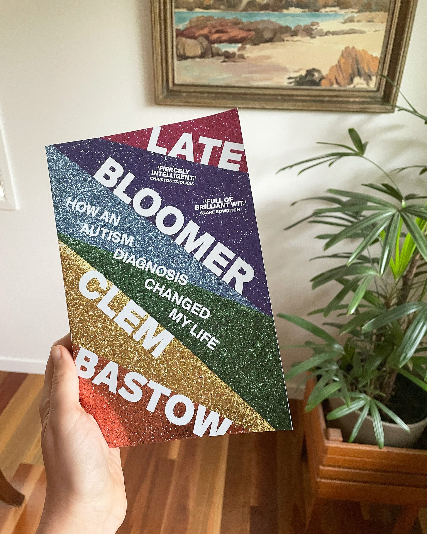 I am holding a copy of 'Late Bloomer' a book by Clem Bastow. My living room is in the background, with a plant and a painting. The book has a rainbow glittery cover