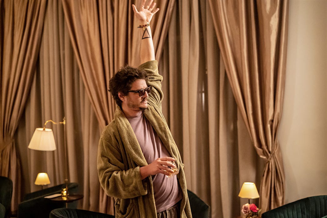 Imagem do filme The Bubble, com o personagem de Pedro Pascal num quarto de hotel, de pijamas e óculos escuros, segurando um copo de uísque, erguendo a mão esquerda, onde é possível ver no pulso uma tatuagem de triângulo