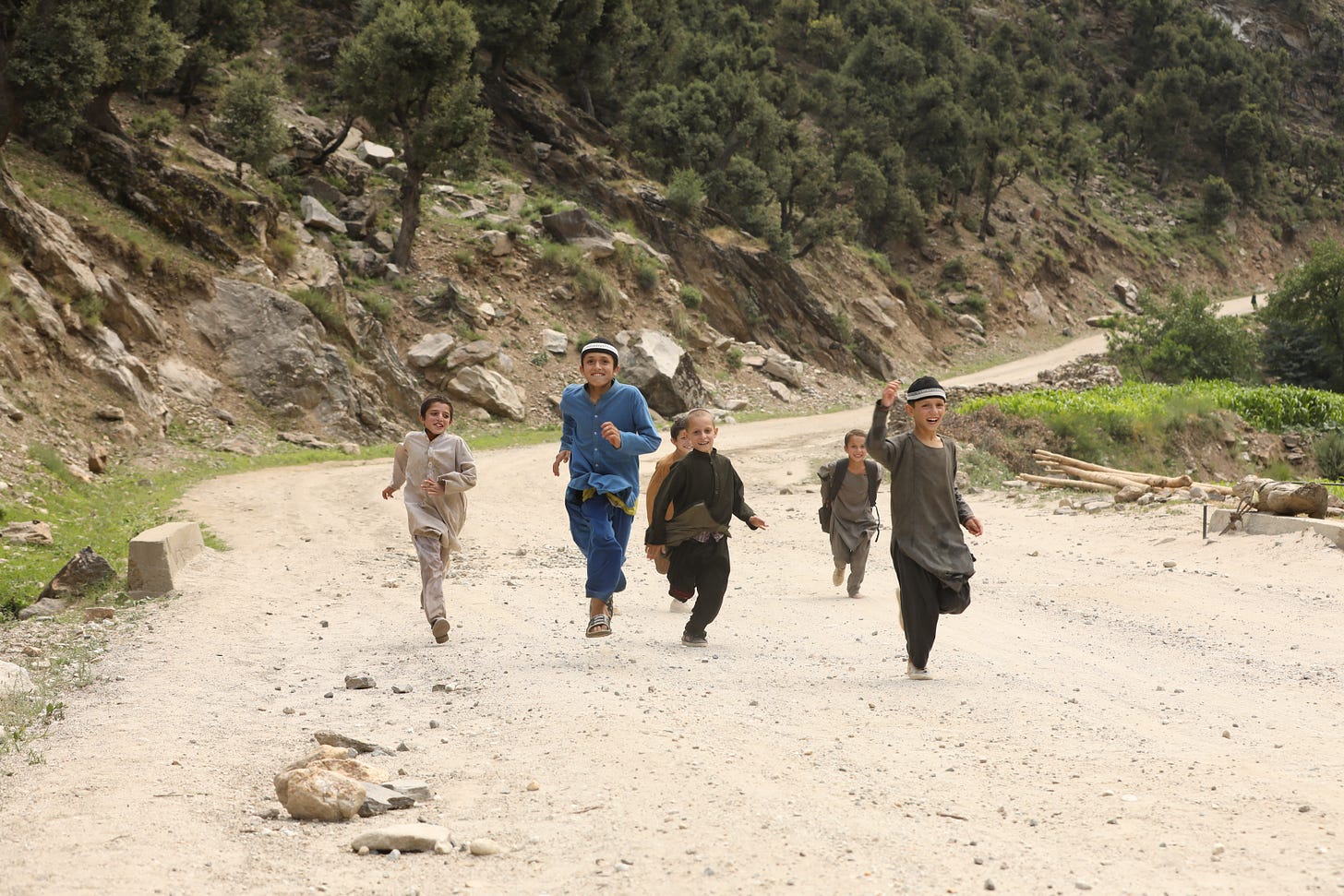 Six smiling children are running down a sandy road in Afghanistan.