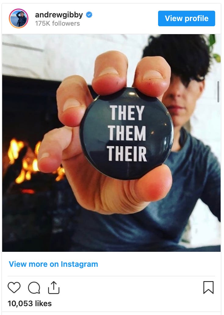 Andrea Gibson showing their pronouns while sitting in front of a fireplace holding a black button with the words "they, them, their" 