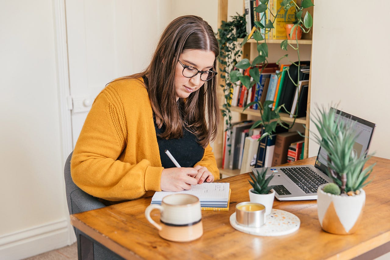 Rachel Baker, freelance copywriter and founder of The Ethical Copywriter, sits at her desk and maps out a content strategy in her notebook. On her desk is a laptop, plants, candle and a mug. She wears an orange cardigan and is gazing down at her notebook.
