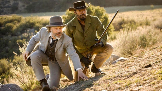 This image shows the bounty-hunting team of Dr. King Schultz (played by Christoph Waltz) and Django Freeman (played by Jamie Foxx) crouching on a hill as they look off-screen at their quarry. Schultz is on the left and Django, who holds a rifle, is on the right. This image shows the bounty-hunting team of Dr. King Schultz (played by Christoph Waltz) and Django Freeman (played by Jamie Foxx) crouching on a hill as they look off-screen at their quarry. Schultz is on the left and Django, who holds a rifle, is on the right.