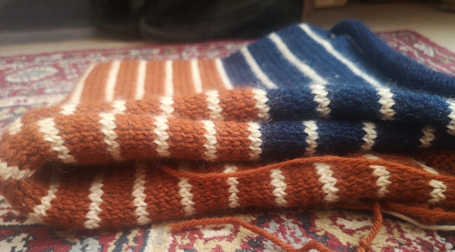 a photo of a large square of knitting, folded over on top of a red moroccan-style rug; the knitting consists of large orange-red rows with smaller white stripes, and at the bottom navy blue rows with white stripes.