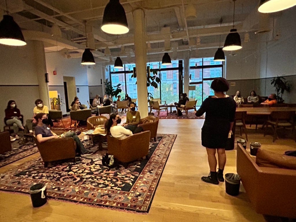 In a wide open room with exposed HVAC, big black hanging lights, a large floor to celing window, brown chairs and wooden tables and printed rugs, Kristen stands addressing a masked group of people on crochet technique