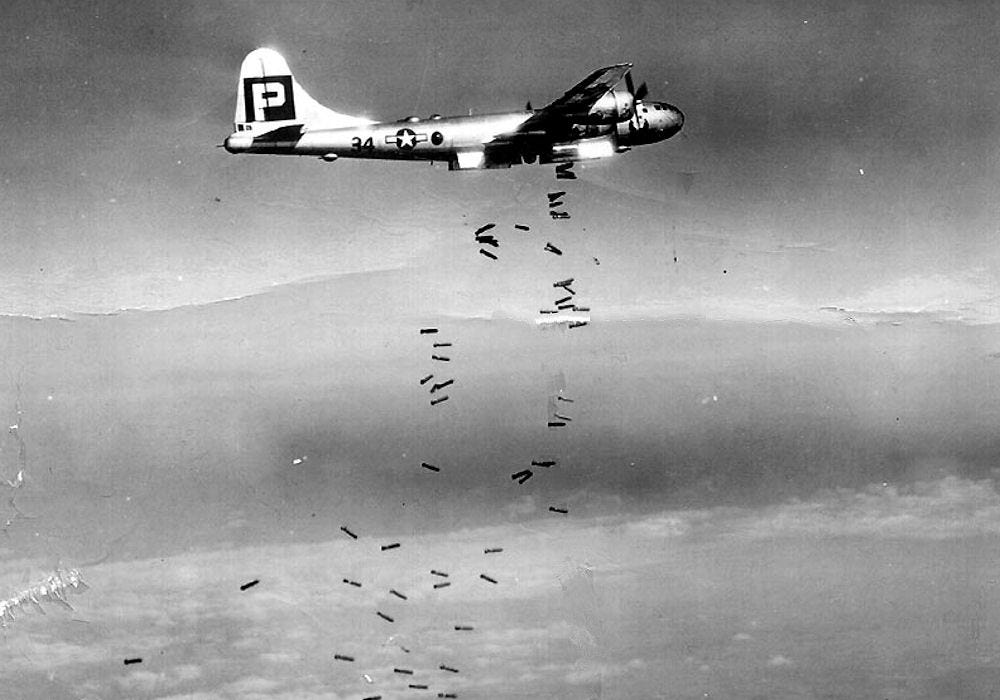 A bomber flies through the sky. Dozens of bombs can be seen dropping from the plane. 