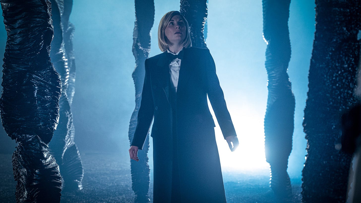 This image shows the perplexed Thirteenth Doctor (played by Jodie Whittaker) standing inside a strange forest, amidst strange, ganglia-like tree trunks. A bright blue light suffuses the image.