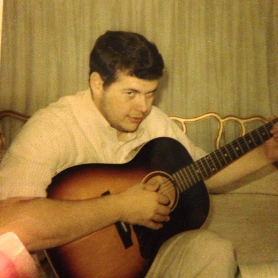 Young white man sitting on a bed with a guitar.