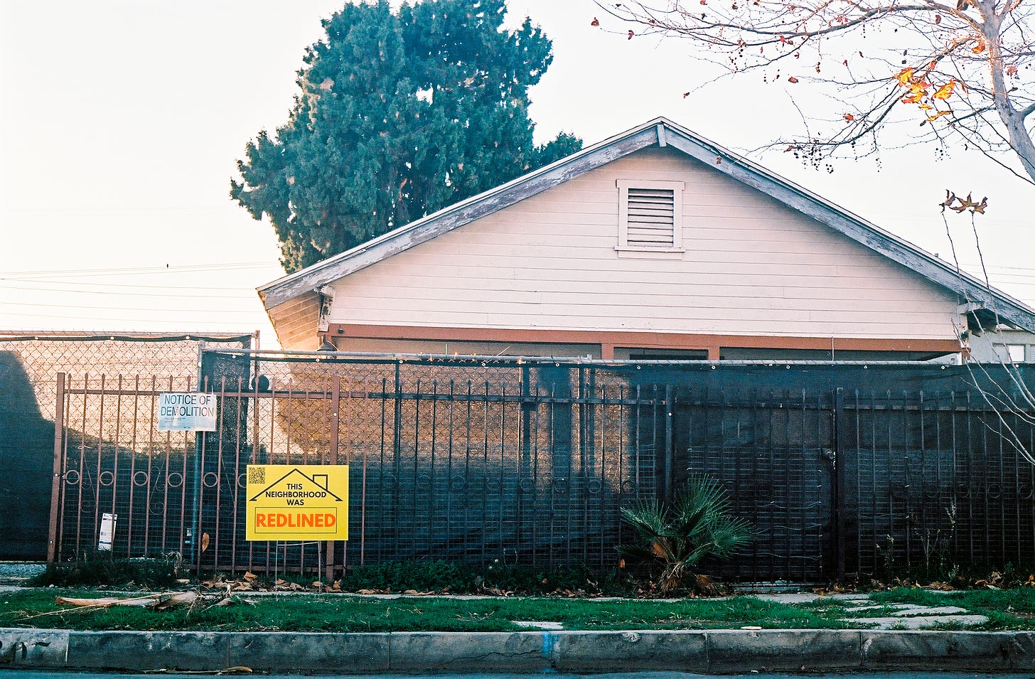 Photograph of a pink house behind a metal fence with a Notice of Demolition sign and one of my yard signs in the parkway in front of the home.